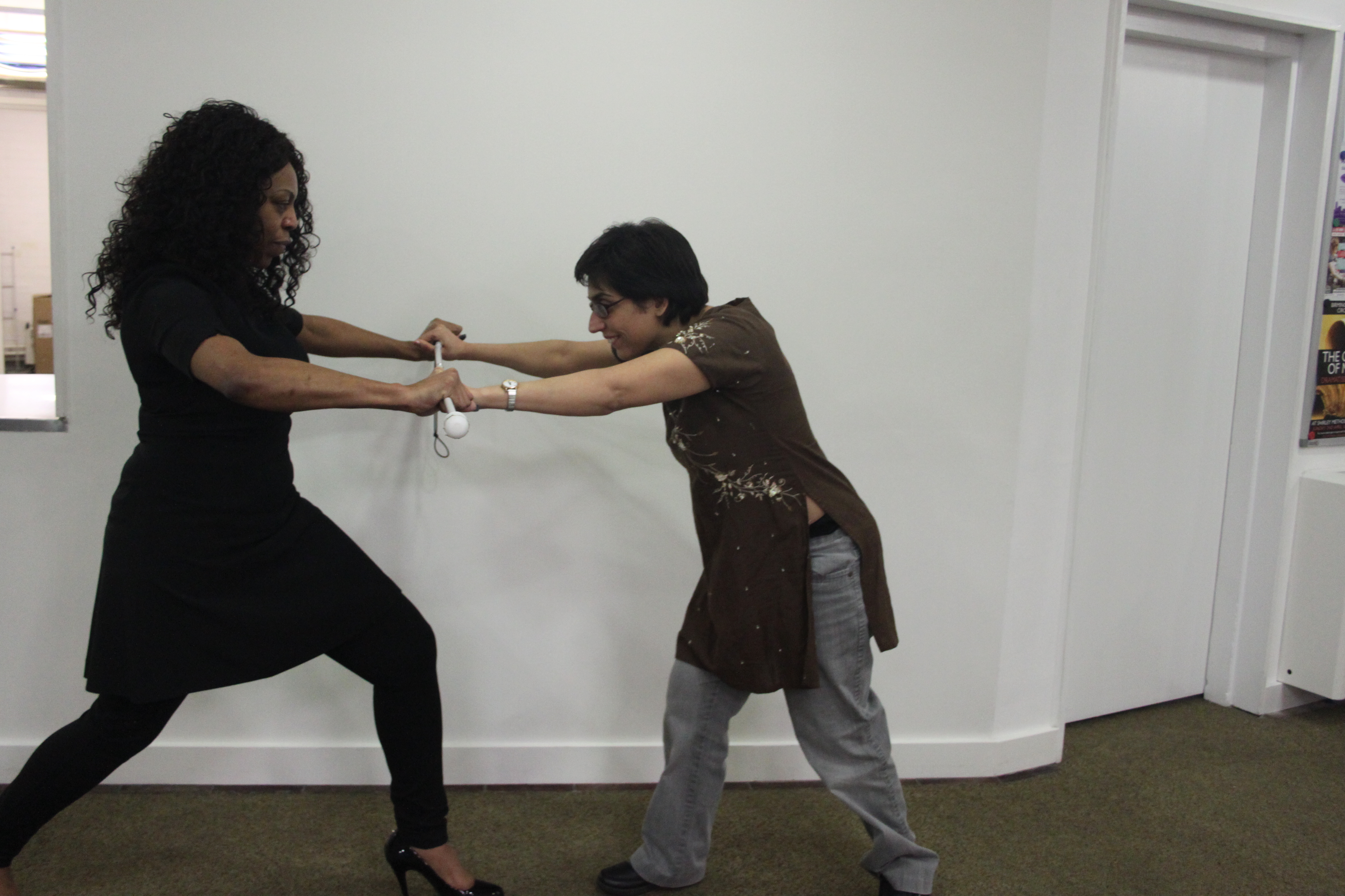 two women facing opposite one another in lunge position, both holding on to a white cane acting as a bar in between them. One resists the push/pull of the other.