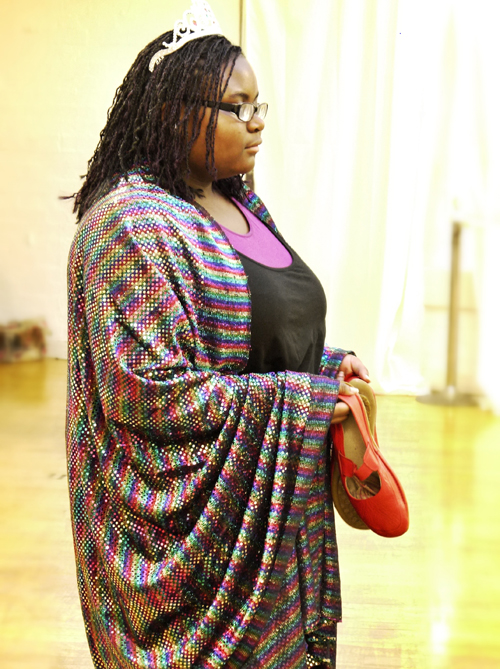 A close up profile shot of the black female actor standing quietly in her technicolour robes holding a pair of red shoes in both hands.