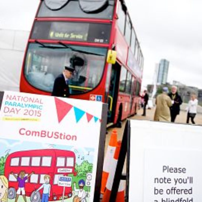 In the foreground a sign for ComBUStion with a cartoon of a red bus and children excited to board. Next to this sign is another saying 'Please note you'll be offered a blindfold.' In the background is the double decker Combustion bus with the conductor and members of the public standing beside it.