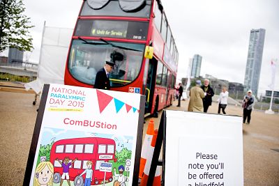 In the foreground a sign for ComBUStion with a cartoon of a red bus and children excited to board. Next to this sign is another saying 'Please note you'll be offered a blindfold.' In the background is the double decker Combustion bus with the conductor and members of the public standing beside it.
