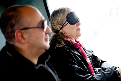 Two passengers, a man and woman, sit next to each other on the bus. The woman is wearing a blindfold and the light from the bus window is reflecting off her face as she tilts her head up.