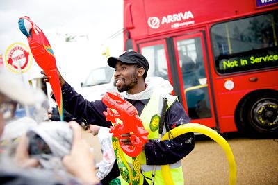 A black male bus attendant holds out an inflatable parrot and lizard to attract passengers' attention. Behind him is the front of the bus and to his left a circular bus stop sign.