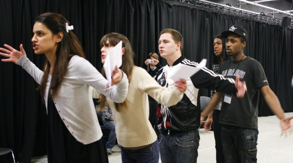 In rehearsal, five young people stand in a diagonal line, holding scripts. Their arms are gestured outwards and their mouths are open, as if talking to someone beyond the camera.
