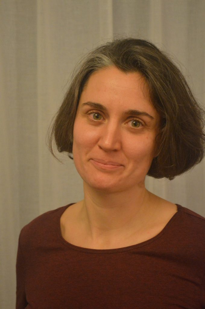 A medium close-up of a white woman with cropped dark hair inside and wearing a maroon top.