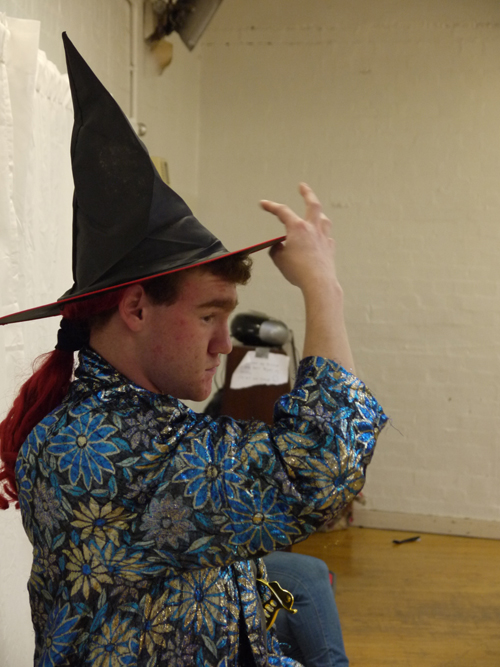 A close up of the male actor sitting at the side of the room adjusting his black wizard hat with his right hand.
