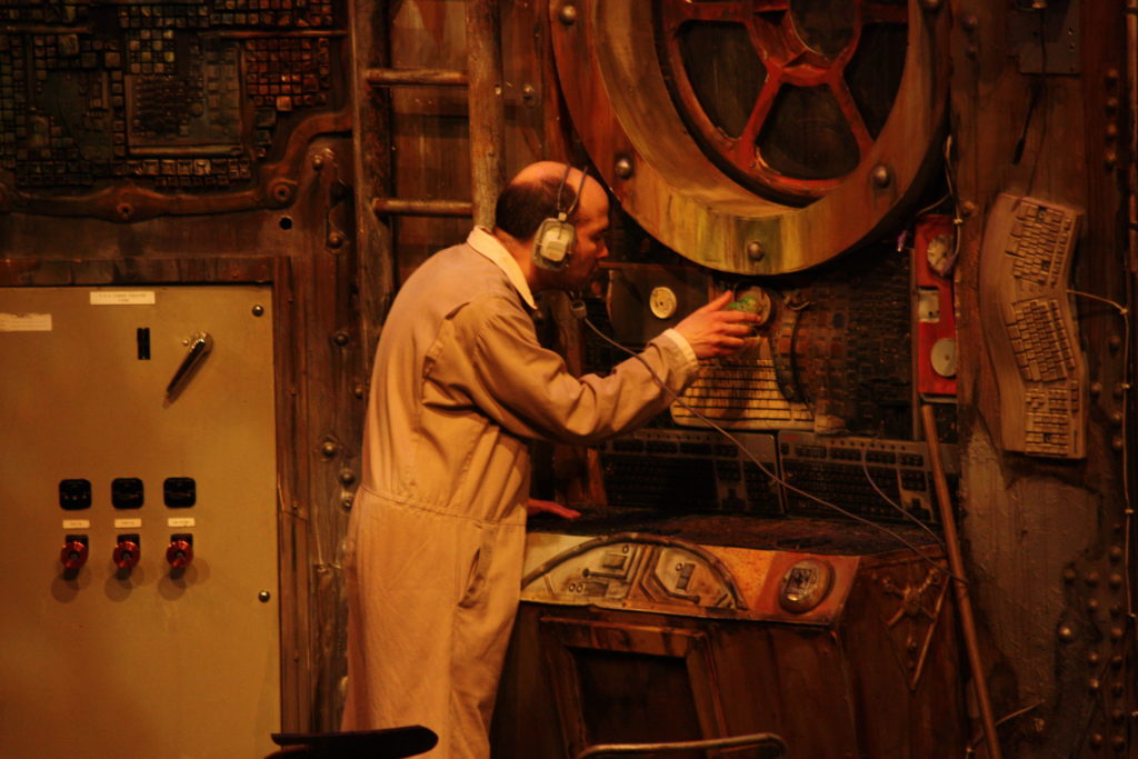 Scene from The Chairs 2016 - Old Man (Tim Gebbels) with headphones on turning a dial on a large control panel as if detecting radar signals