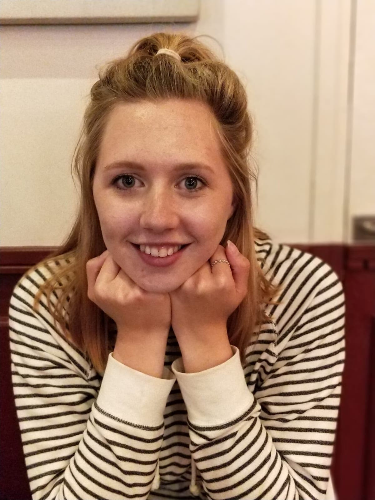 A young woman with blonde, her face resting in her hands, her elbows resting on a table out of sight. she wears a long-sleeved white top with thin black stripes.