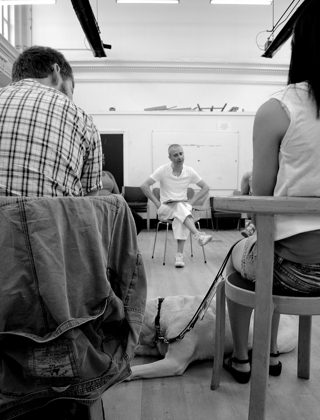 Two participants sit with their backs to us, a guide dog sits between them. They are facing the drama facilitator who can be seen between them, seated opposite talking thoughtfully. Black and white photo