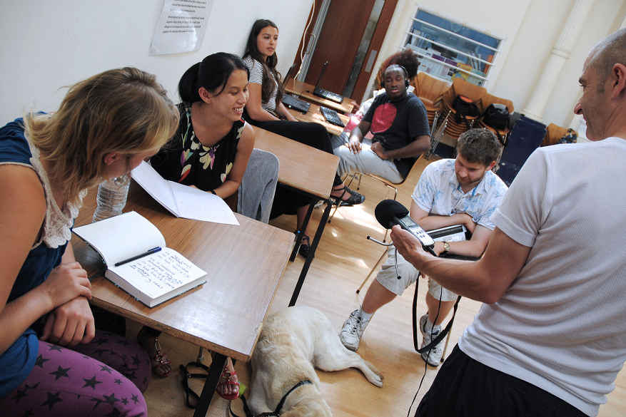 Participants are sat at desks with laptop, books and other writing devices before them, they are smiling at drama facilitator Oli who holds a microphone out towards them