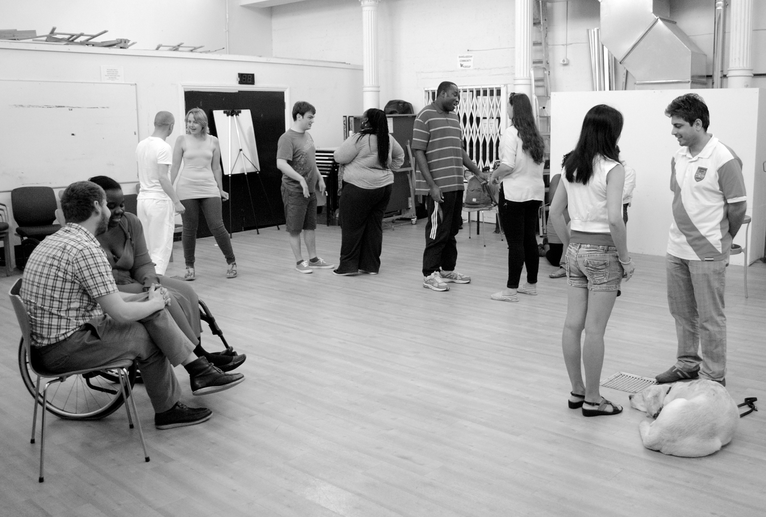 A group of participants are arranged in a semi circle, sitting or standing in pairs they are chatting animatedly. A guide dog is curled up next to a woman on the right