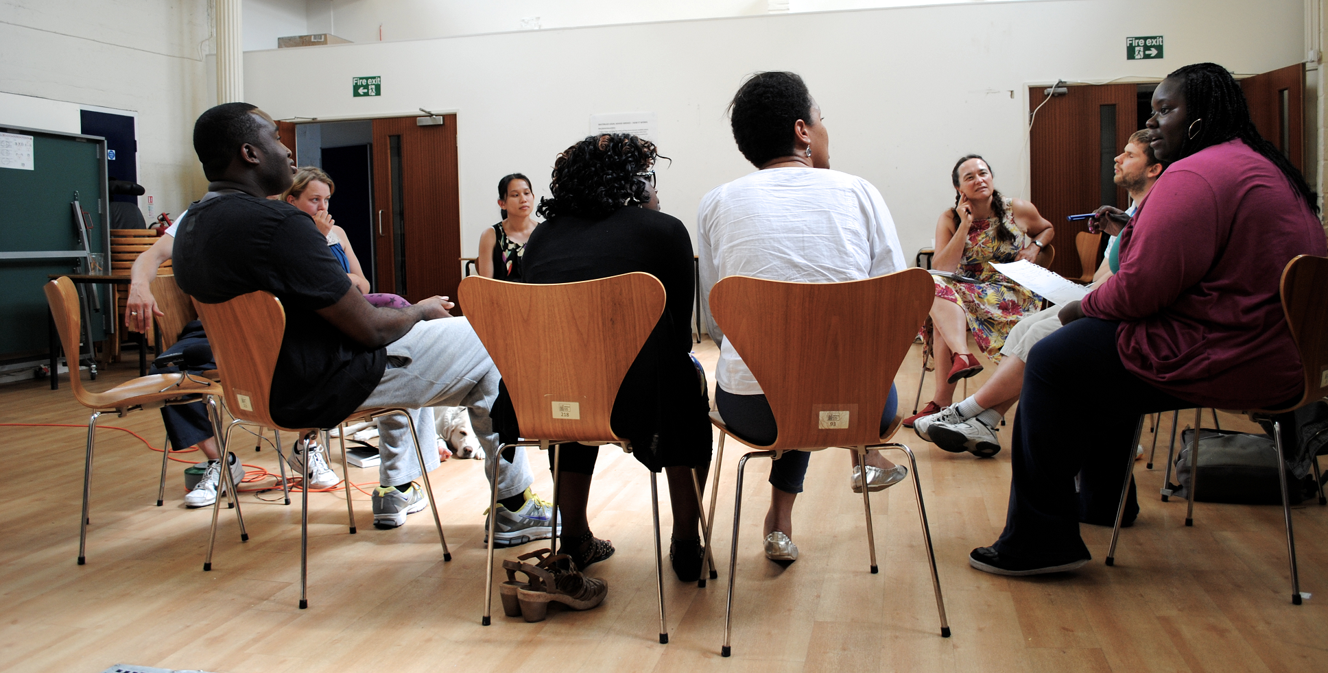 The view from outside a circle of chairs, the workshop group are listening to a woman talking on the right