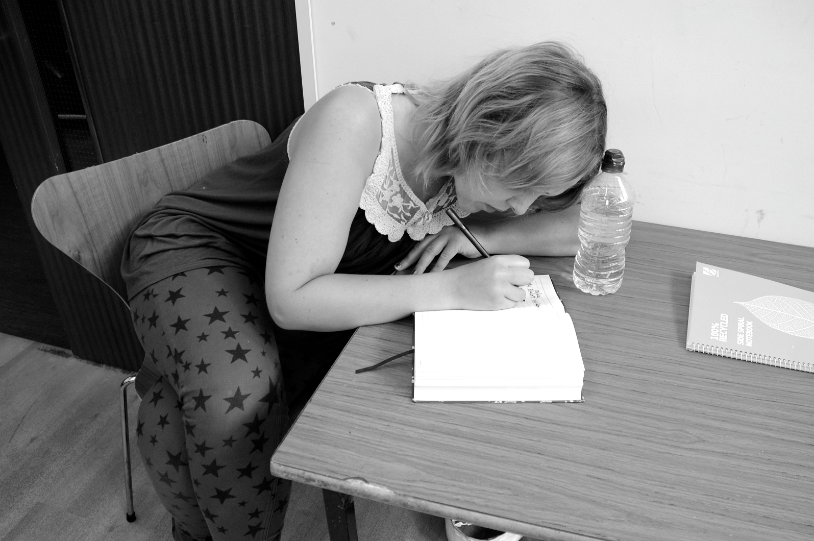 A white woman leans on a desk in concentration as she writes in a notebook. Black and white image