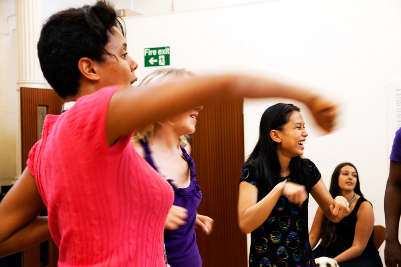 Three young women are taking part in a physical warm up, making punching movements as one giggles. An access worker looks on