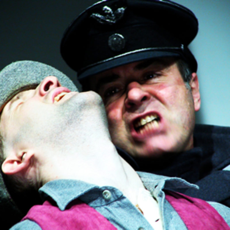 A close up image of white man grimacing in a policeman's hat and coat pulling back the head of another white man in a flat cap and a grey shirt.
