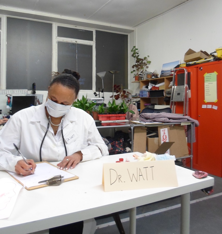 A woman in doctor's outfit and face mask sits busy behind a desk on which a sign reads Dr Watt