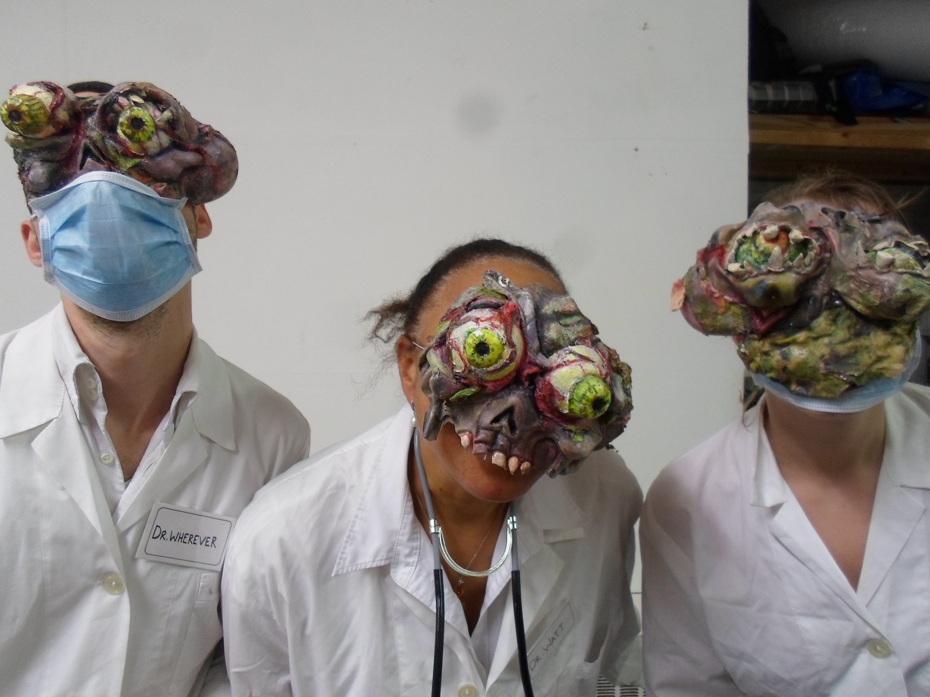 Three people in white coats and face masks lean towards the camera, each wears grotesque prosthetic eyes 