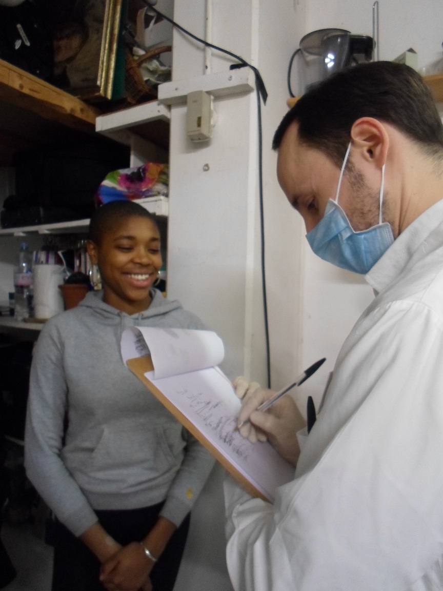 A white man wearing a doctor's uniform and face mask writes down notes on a clipboard while a smiling black woman stands to the side.