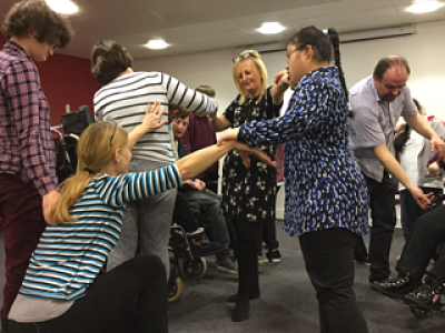 A group of workshop participants stand and crouch in different positions while linking arms with one another.