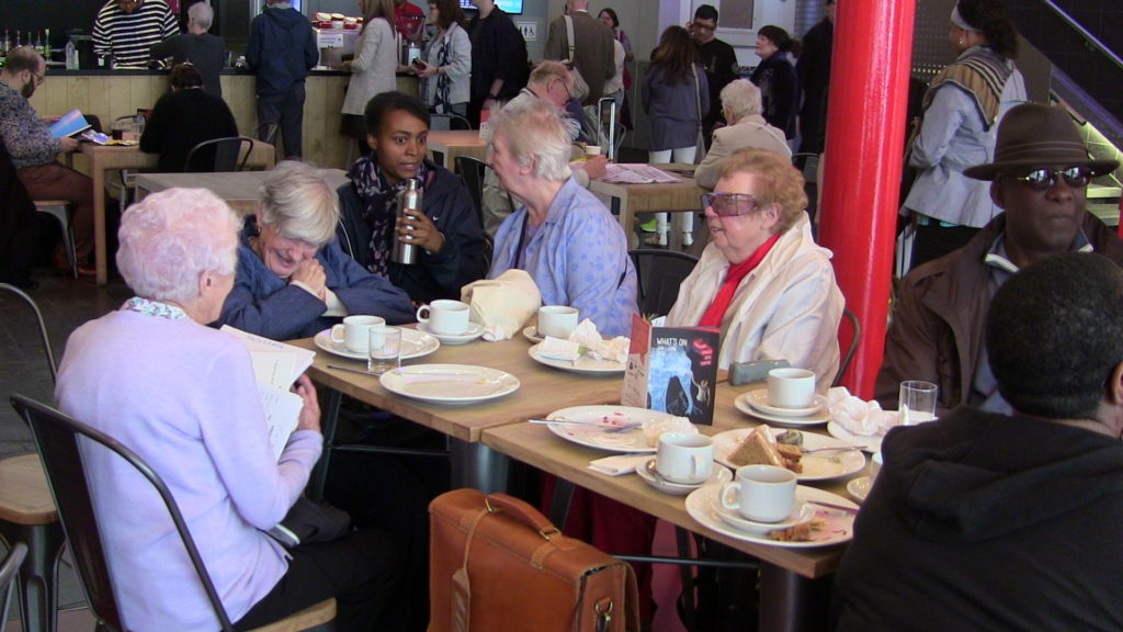 A group of older women sit around a table in the foyer at Stratford Arts Centre talking to a younger access worker.
