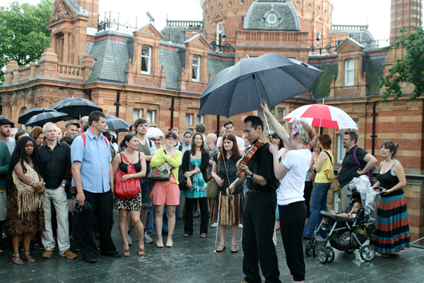 Japanese viola player with audience around him and umbrella over him. Film of an outdoor performance piece