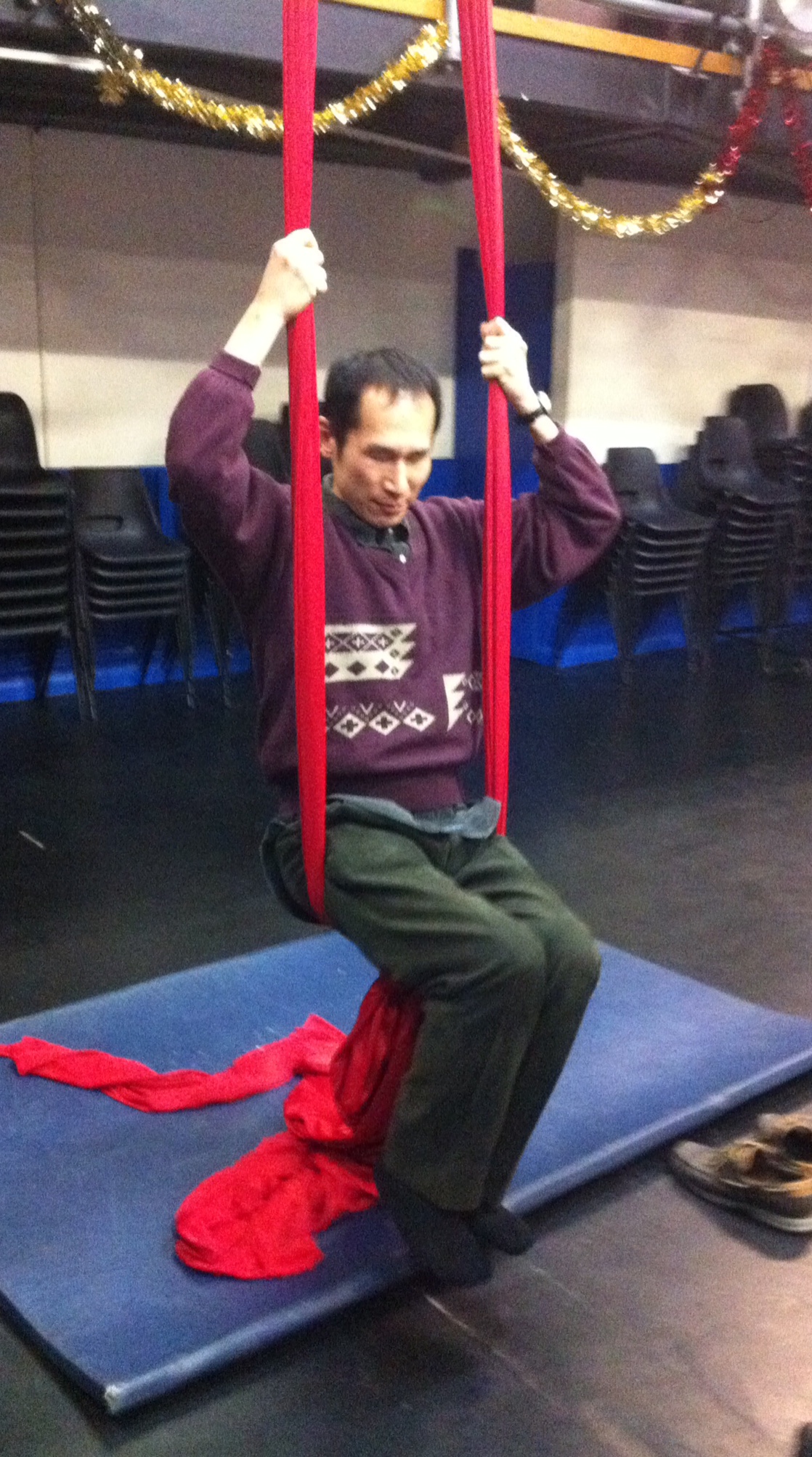 Takashi, a Japanese man, sits on a swing made of red silk material, holding on with his hands