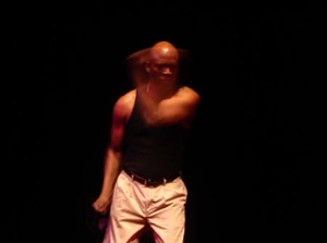 A black male performer standing on stage, his left arm motioning across the stage in a fast blur.