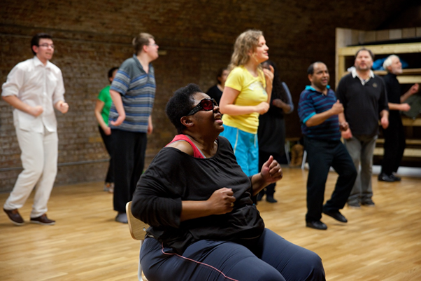 Mixed group of participants in lines, copying a marching movement, a black woman in the foreground is doing the movement seated