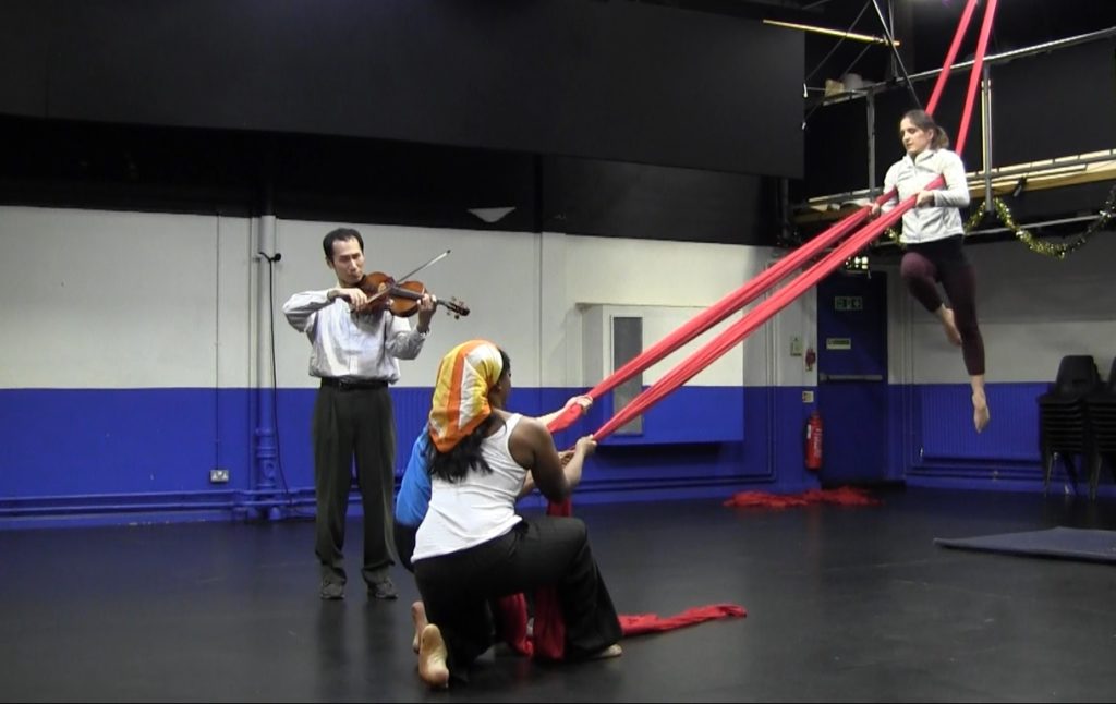 A white female aerialist hangs from two bolts of red silks strung from the ceiling. A Japanese viola player is playing