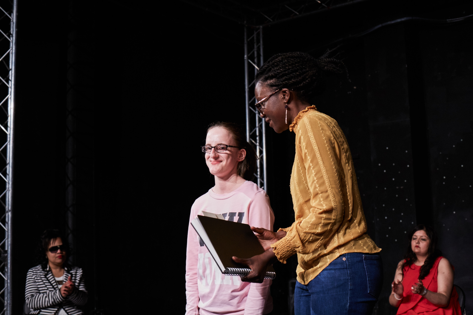 Emily and project manager Stella, who holds Emily's notepad from which she reads out her poem. Two female performers in the background applaud her reading.