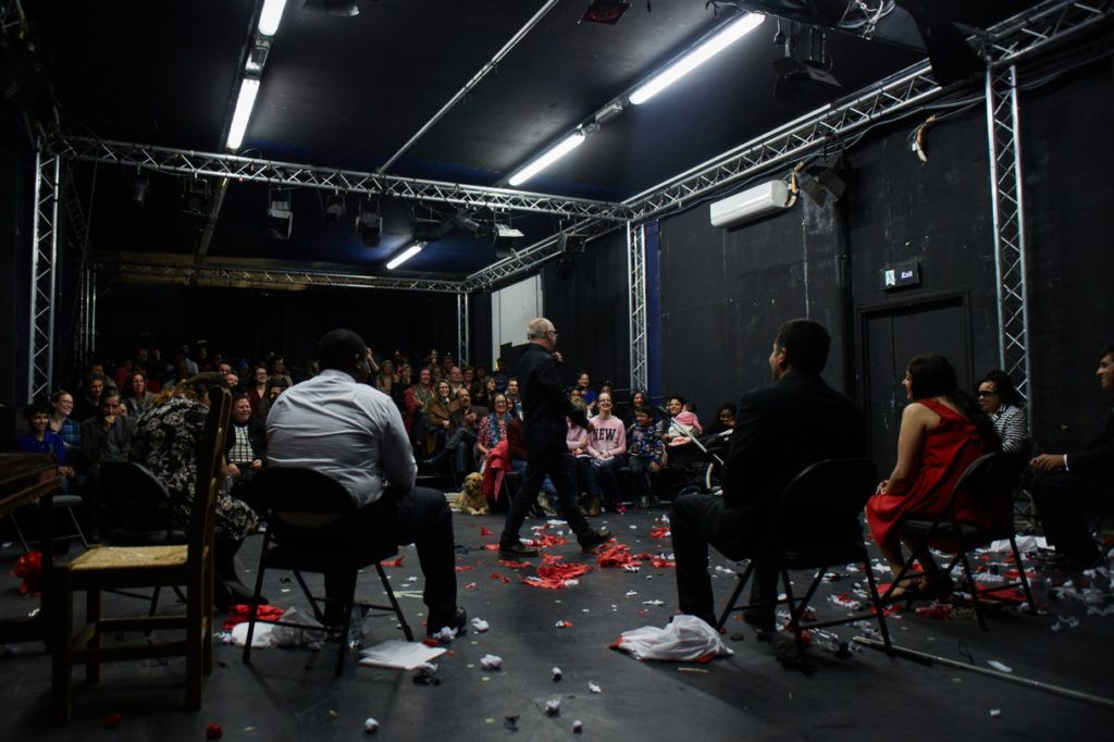 Shot from behind of Unexpected participants in chairs sitting in front of audience and director Shane walking across the stage