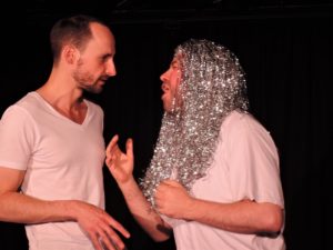 A close up shot of two white men standing facing each other. One wears a long silver tinsel wig and the other looks at him, with a confused expression.