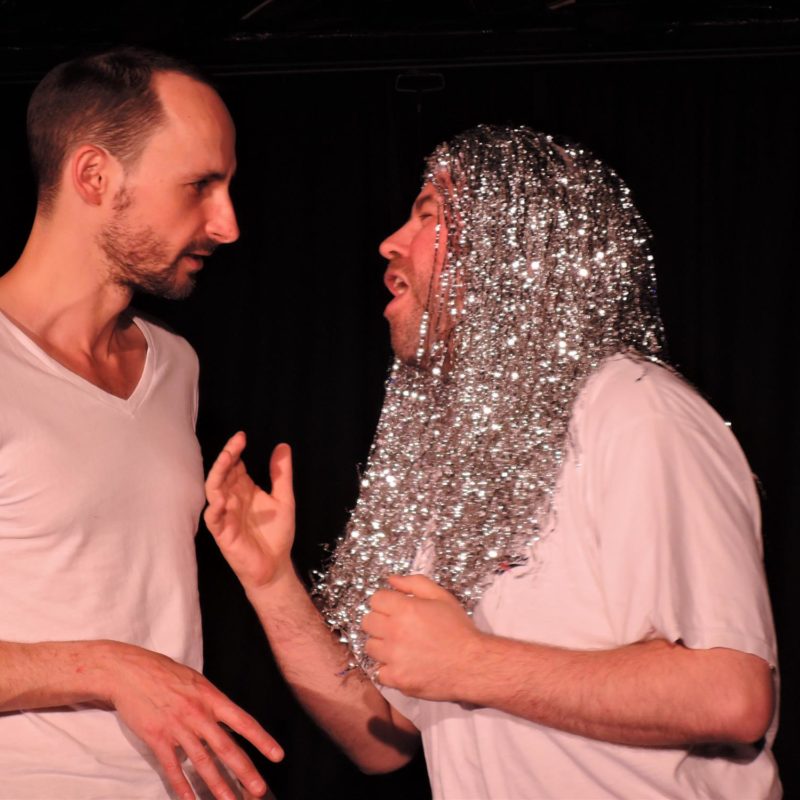 A close up shot of two white men standing facing each other. One wears a long silver tinsel wig and the other looks at him, with a confused expression.