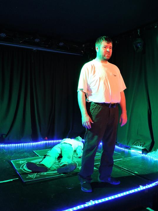 A white man stands on a stage, lit by a green light. He looks out towards the audience. Behind him, a tall white man lays on the floor. 