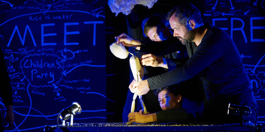 Three puppeteers dressed in black hold a neutral human puppet, which looks down to the ground. A small light is projected onto the puppet.