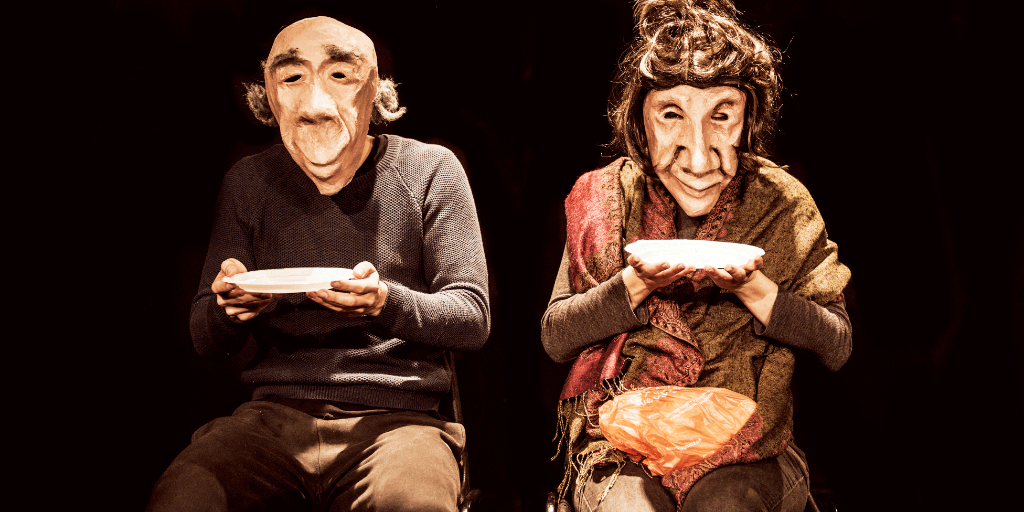 Elderly masked male/female couple sitting side by side holding plates.