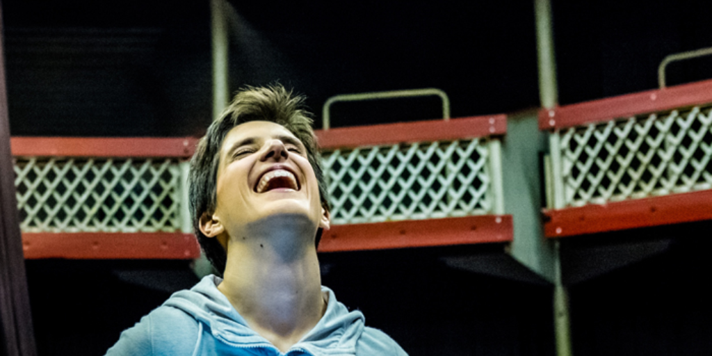 A mid shot of a young woman throwing her head back and laughing. She is onstage in the auditorium of a small-scale theatre.