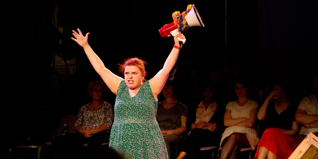 A young woman performing in-the-round. Her arms are raised and she carries a megaphone.