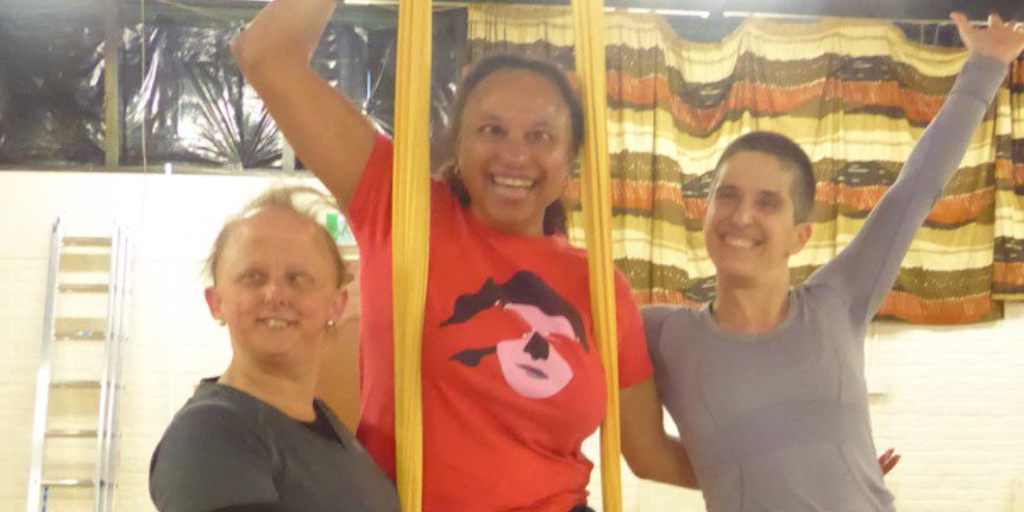 A mixed race woman in a red t-shirt supported on yellow aerial silks by two white women. The woman to the left has thin