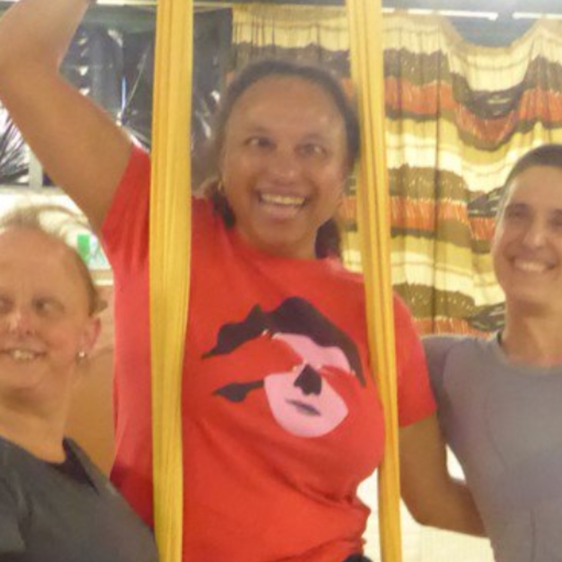 A mixed race woman in a red t-shirt supported on yellow aerial silks by two white women. The woman to the left has thin