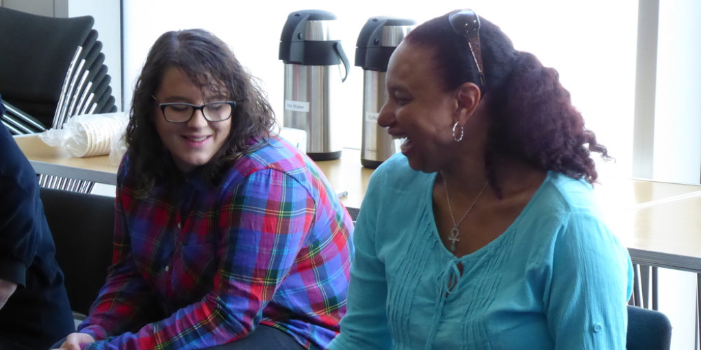 A medium shot of a seated young white woman with dark hair and glasses and a seated mixed-race woman with sunglasses on her head. They are i