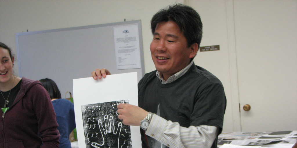 A Japanese man wearing a shirt and green jumper holds up a textured image of a hand.