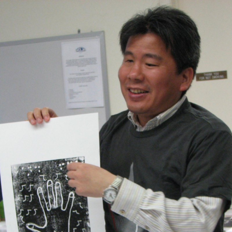 A Japanese man wearing a shirt and green jumper holds up a textured image of a hand.