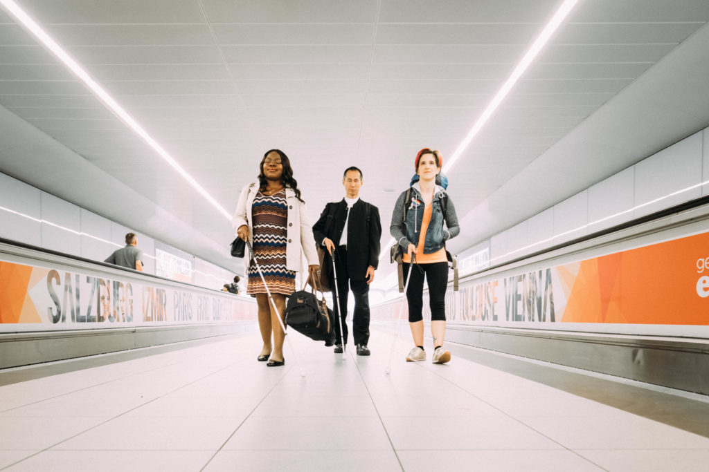 A landscape photo of Victoria, a Nigerian woman, Takashi, a Japanese man and Amelia, an Italian American woman. The three stand in a line, walking towards the camera between two airport moving walkways