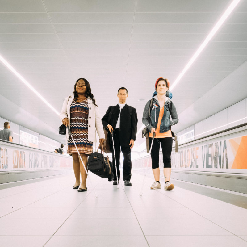 A landscape photo of Victoria, a Nigerian woman, Takashi, a Japanese man and Amelia, an Italian American woman. The three stand in a line, walking towards the camera between two airport moving walkways