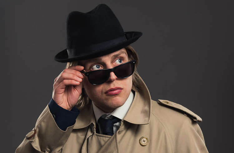 Tom Skelton looking over dark glasses dressed in a beige trench and black trilby