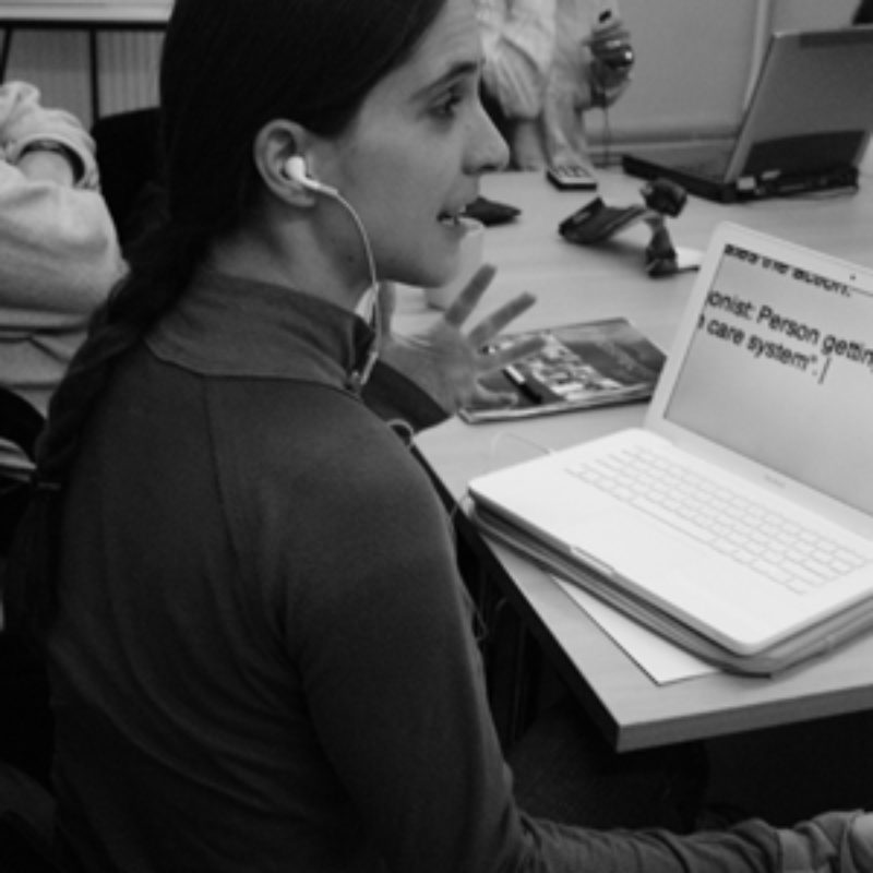 Black and white image of a woman sitting at her laptop (on desk). On the screen is text in large font. She is gesturing with her left hand