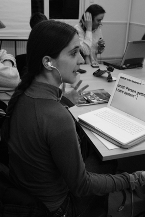 Black and white image of a woman sitting at her laptop (on desk). On the screen is text in large font. She is gesturing with her left hand