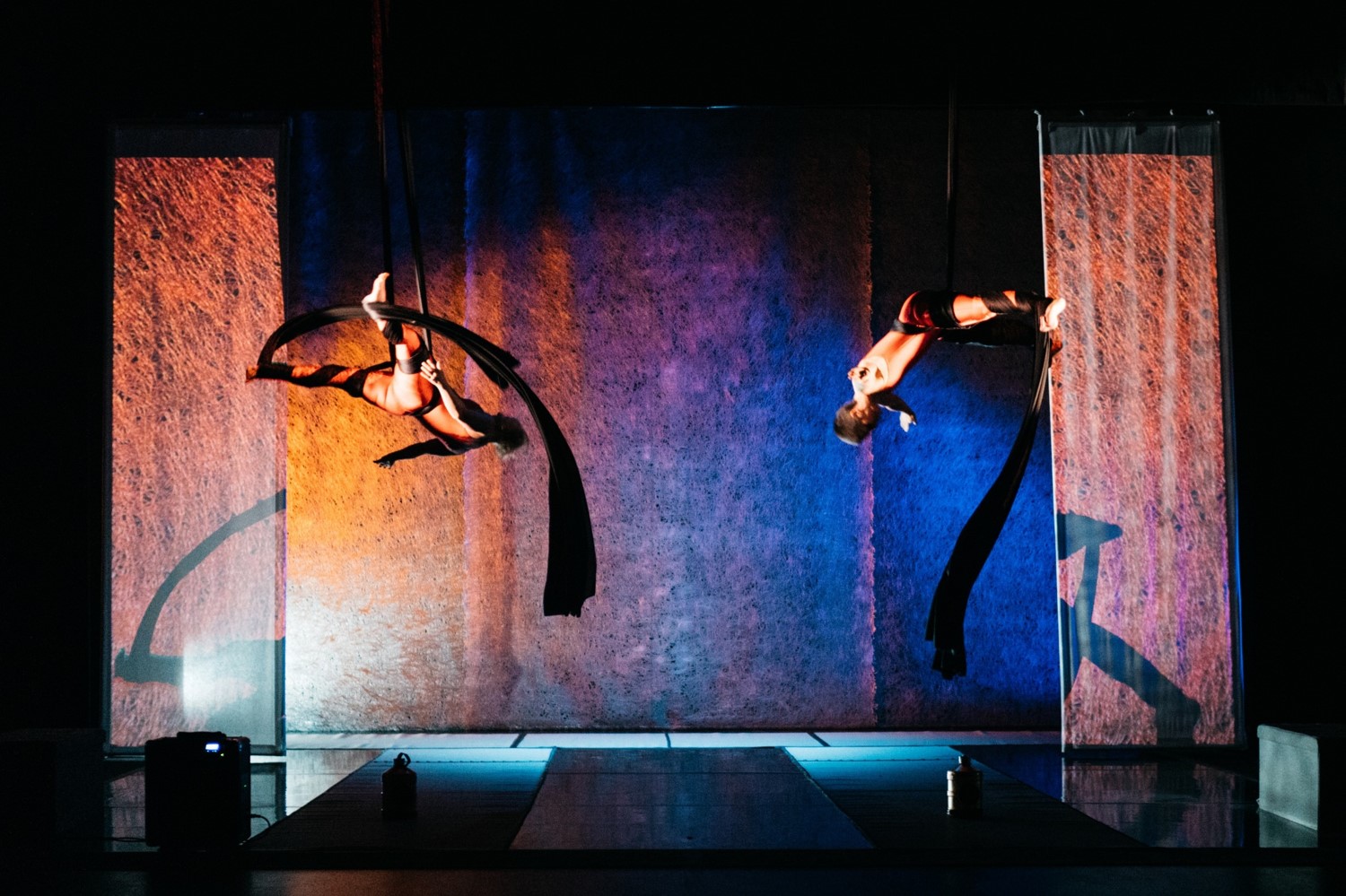 Flight Paths - A wide landscape shot of two women on aerial silks, tumbling through the air mid fall amongst a sea of orange and blue stage lighting.