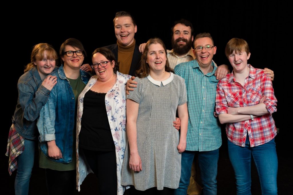 The Pathways Actors, a group of three men and five women, stand together. Their arms around on another they smile and laugh at the camera.