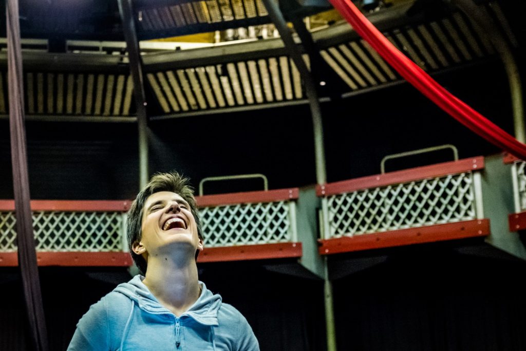 Flight Paths R&D - A medium close up of a woman with cropped hair throwing her head back in laughter. She wears a sky blue, zip up hoodie. She is in a theatre auditorium and aerial silks are visible behind and above her.